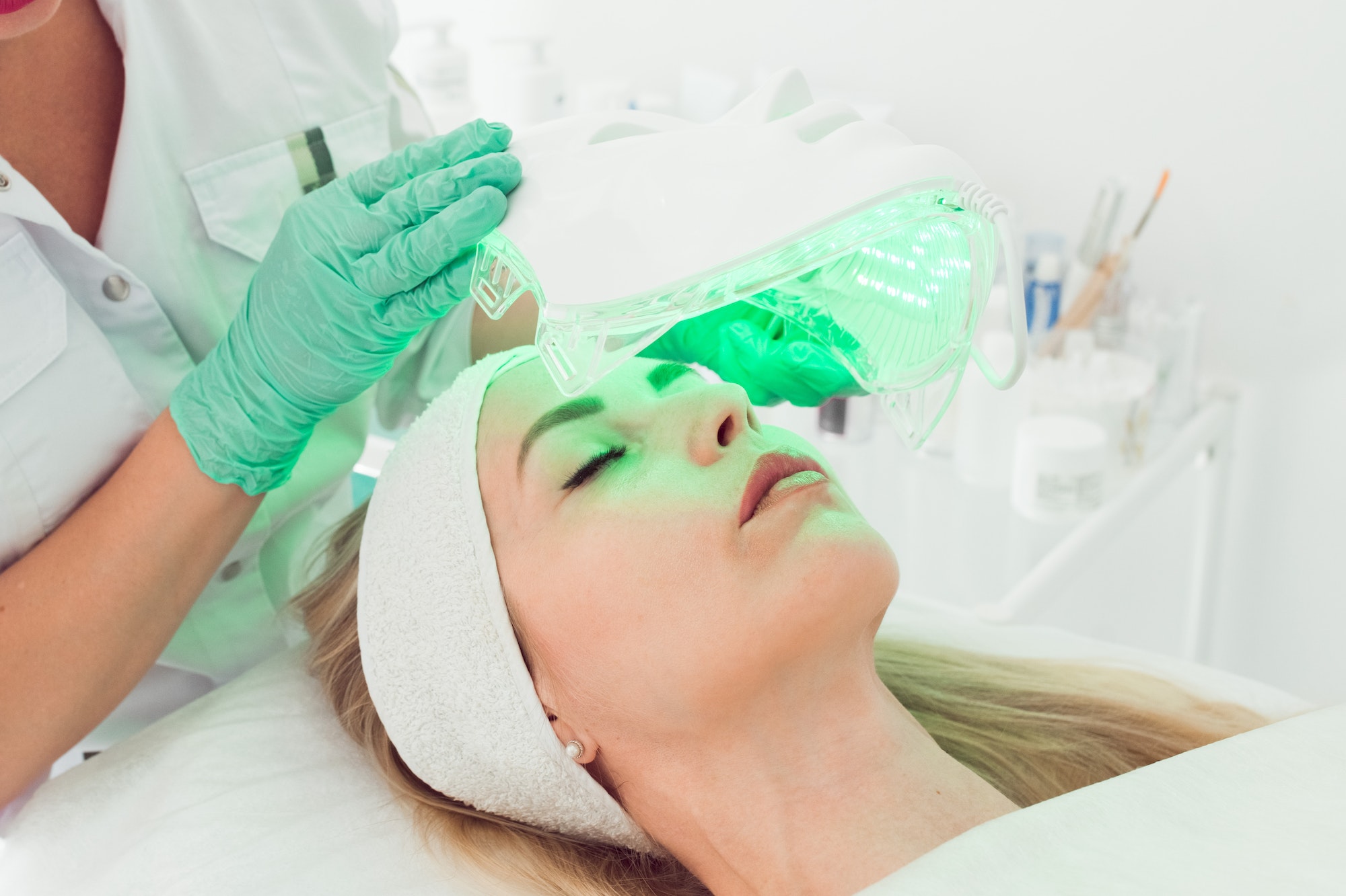 cosmetologist applying light therapy LED facial mask to woman at esthetic clinic